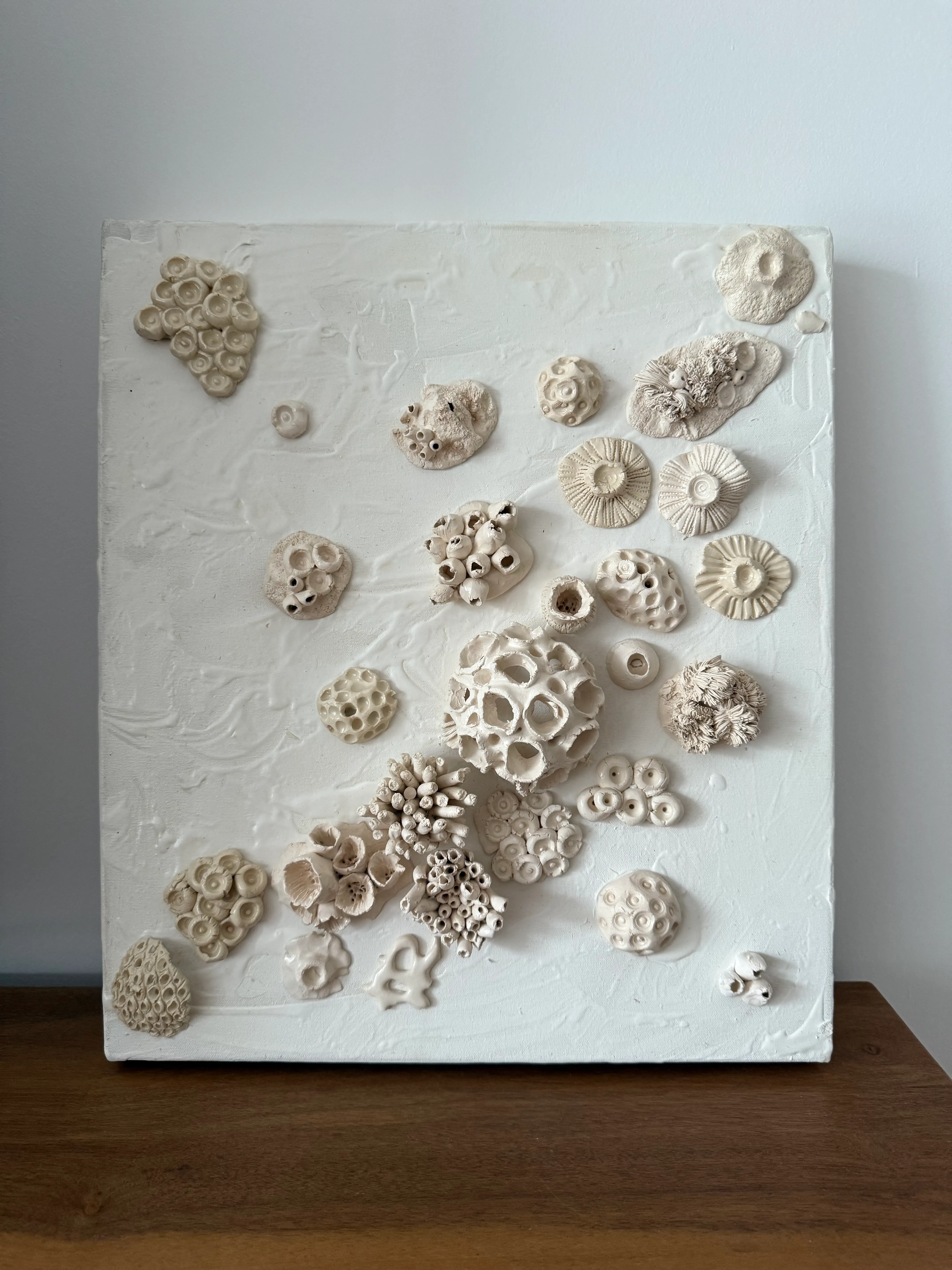 Canvas leaning on a wooden surface against a pale wall. Clusters of white ceramic rosettes, rings and pierced forms spread across a heavily textured white ground.