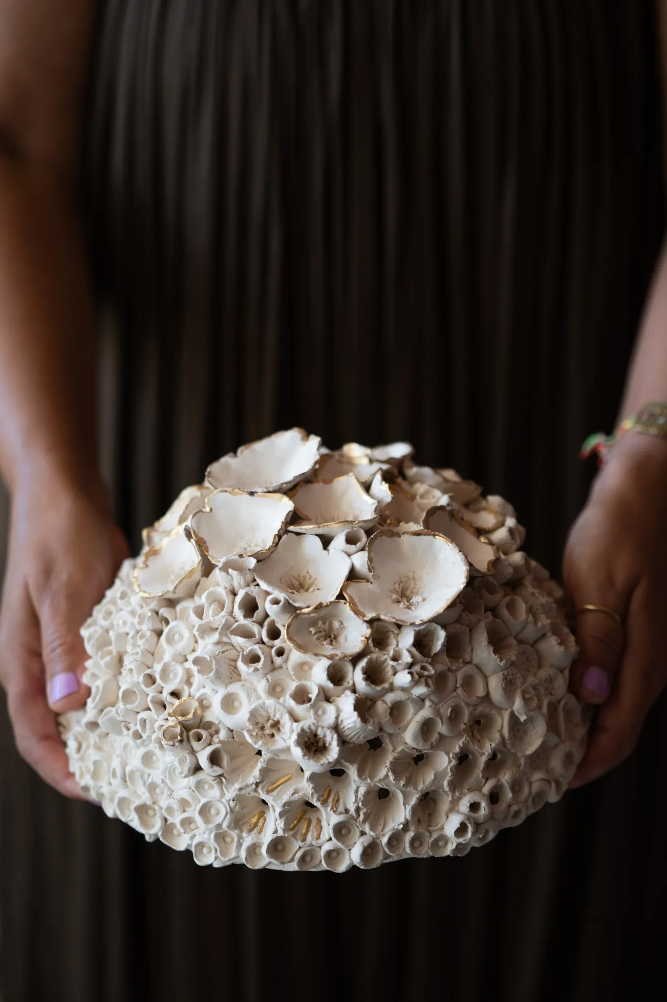 Domed ceramic reef form held in two hands against a dark olive dress. Open tubes cover the lower half, cupped shells gather over the crown.