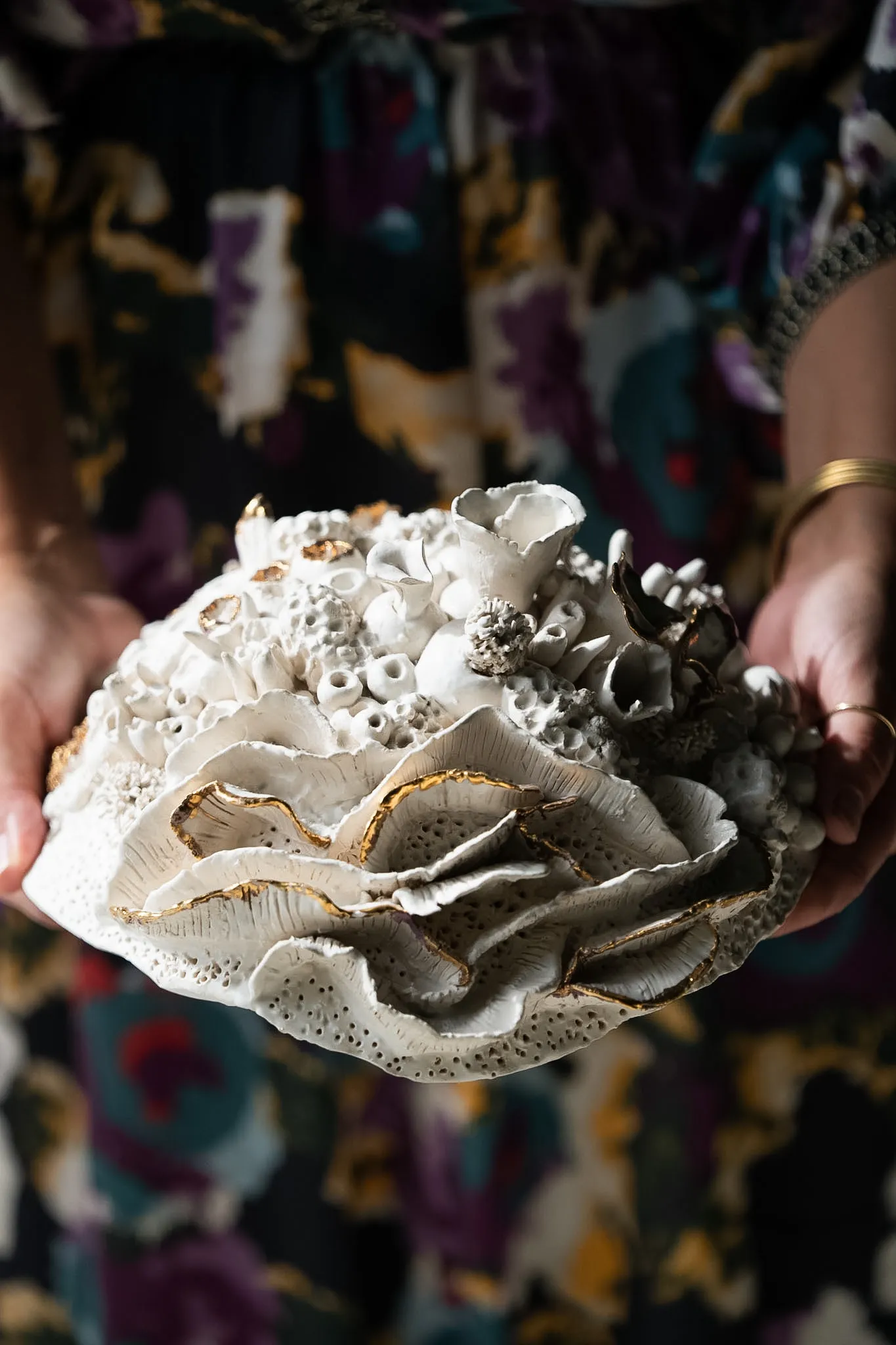 Shallow ruffled ceramic reef form held in two hands against a floral dress. Its wavy edges are rimmed in gold luster above a bed of small tubes.