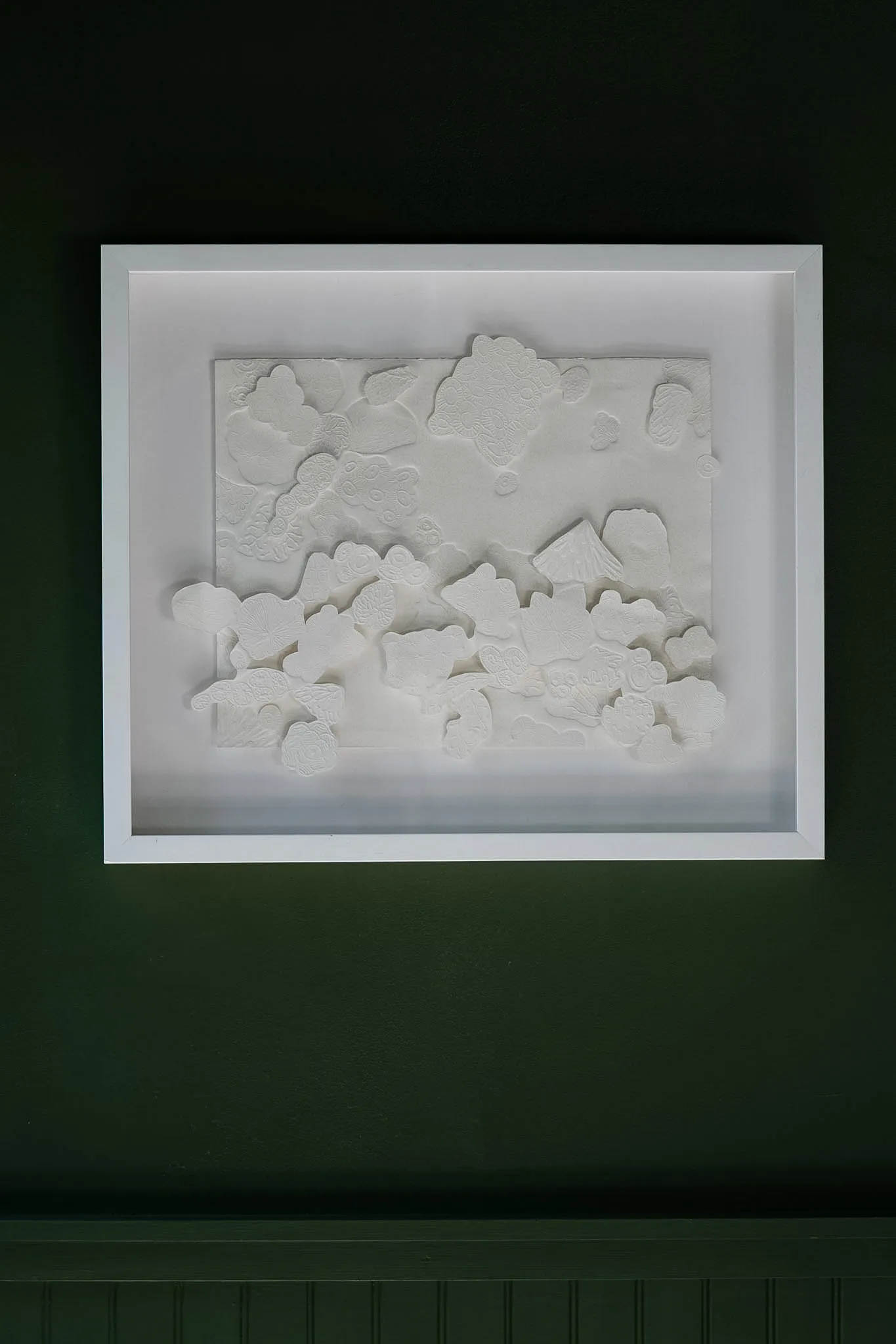 White embossed cotton paper relief in a landscape white box frame on a dark green wall. Layered cut shapes mass along the lower half like a bank of cloud or coral.