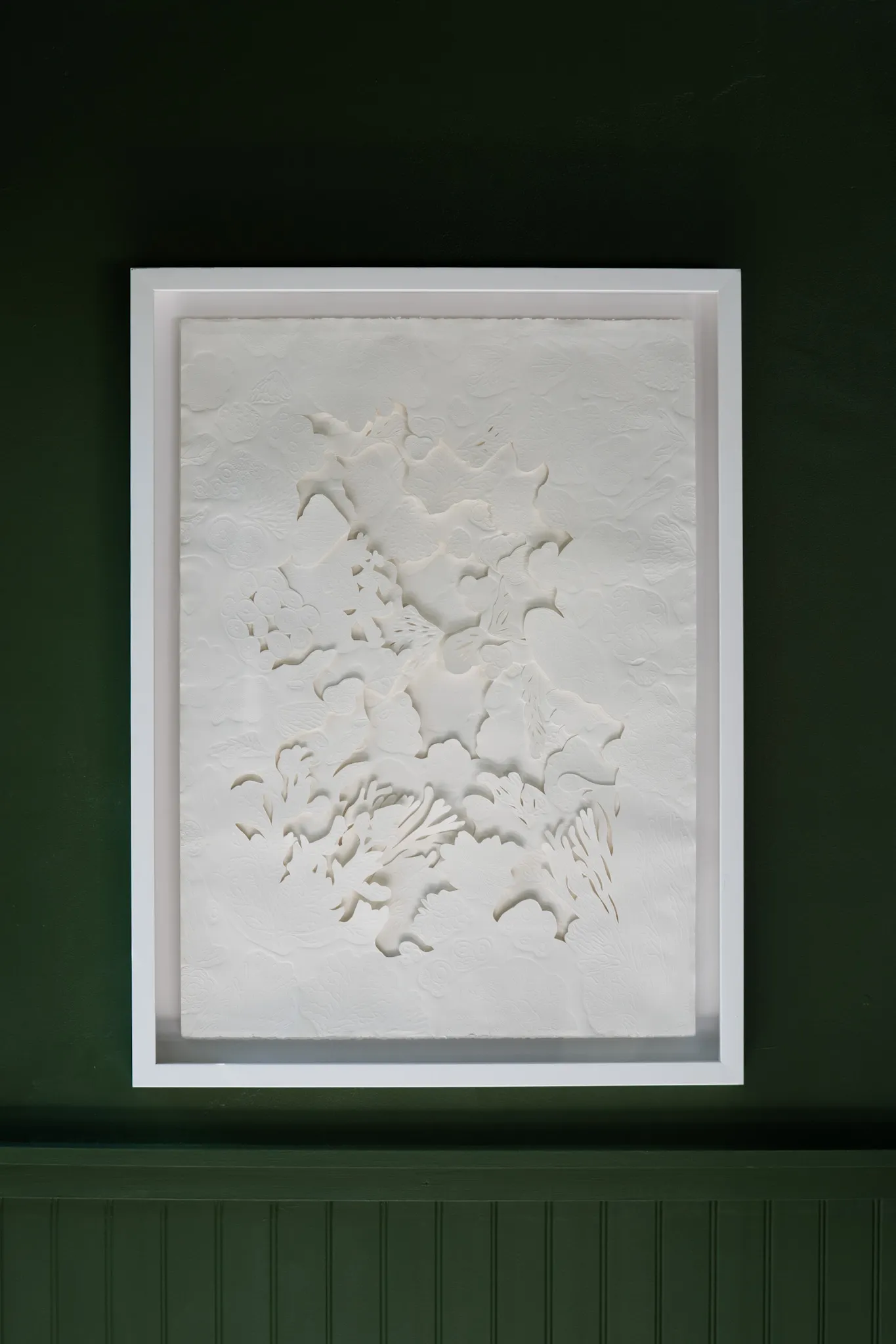 White embossed and hand-cut cotton paper relief in a white box frame, hung on a dark green panelled wall. Ridged forms drift diagonally across the sheet in shallow relief.